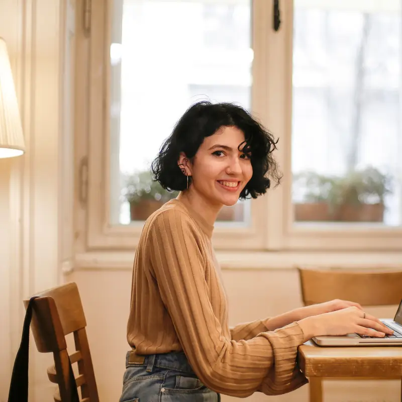Eine junge Frau mit dunklen Locken sitzt am Laptop in einem Café und lächelt selbstbewusst in die Kamera.